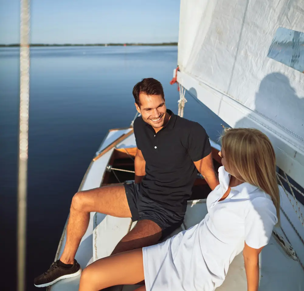 Smiling couple relax on a sailboat deck in calm blue water, leaning together beneath white sails on a sunny summer day.