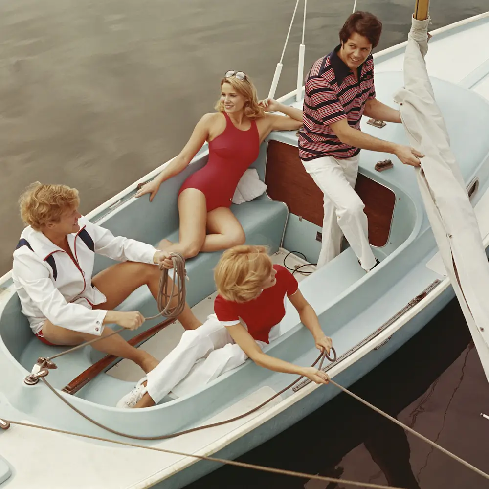 Four cheerful adults relax on a sailboat: a woman in a red swimsuit reclines while others handle ropes on the water.