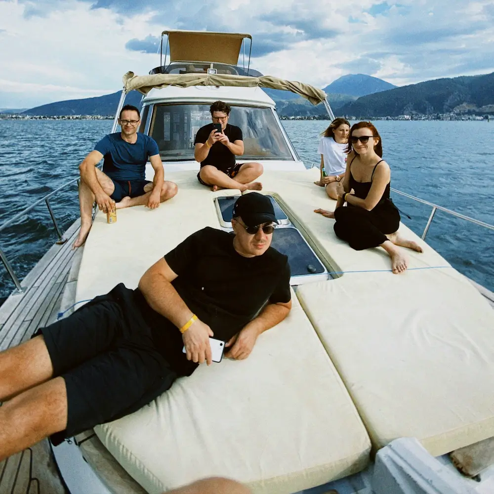 Friends relax on a yacht in a sunny lake, with one man reclining in front and four others seated behind, surrounded by mountains.
