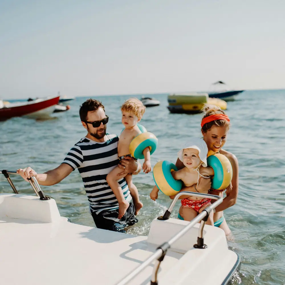 A cheerful family boards a small white boat at sea; the father carries a boy while the mother steadies a baby in colorful swim rings.
