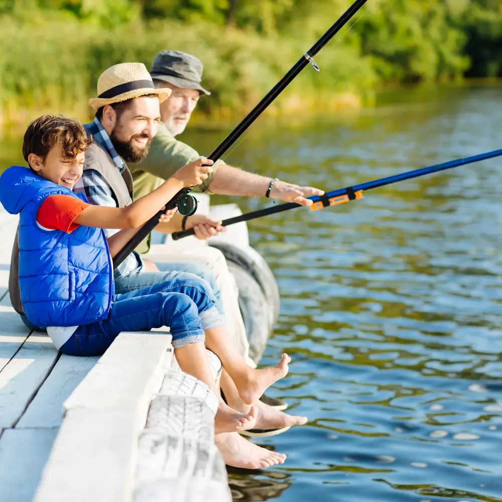 A smiling boy and two adults sit barefoot on a sunny dock, fishing together with rods over a calm lake in a joyful family outing.