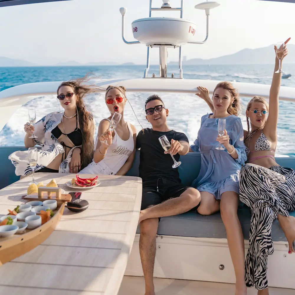 Five friends relax on a yacht, raising champagne glasses with smiles as they celebrate against a bright blue sea and mountains.