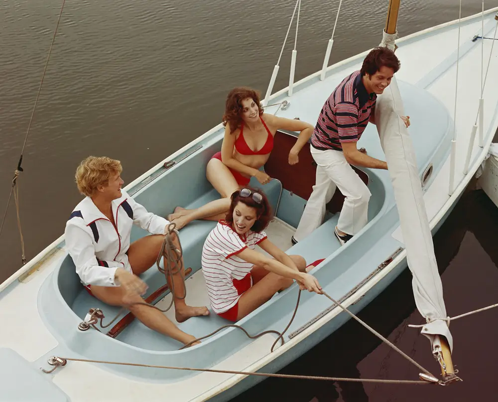 Four young adults in colorful clothes enjoy a carefree sailboat ride, seated among ropes and sails on a calm river.