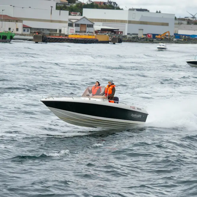 Two people in orange life jackets speed across choppy harbor water in a motorboat, with industrial buildings and boats in the background.