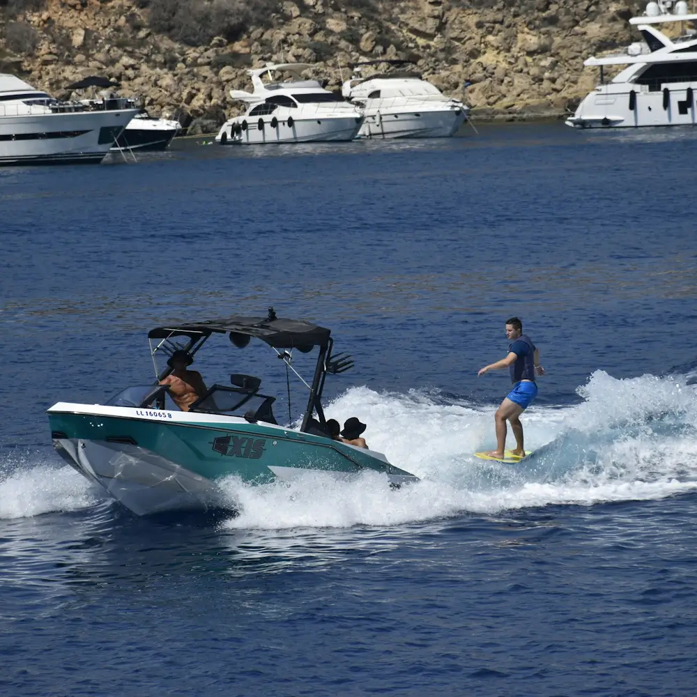 A wakeboarder rides behind a motorboat on blue water, with white spray, anchored yachts, and a rocky shoreline in the background.